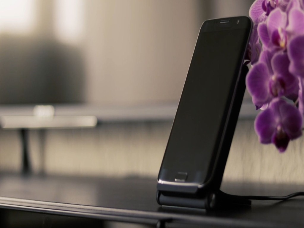 黒いスマートフォンがスタンドに立てかけられ、背景には紫の花が置かれています。シンプルなデザインで、モダンな雰囲気を醸し出しています。