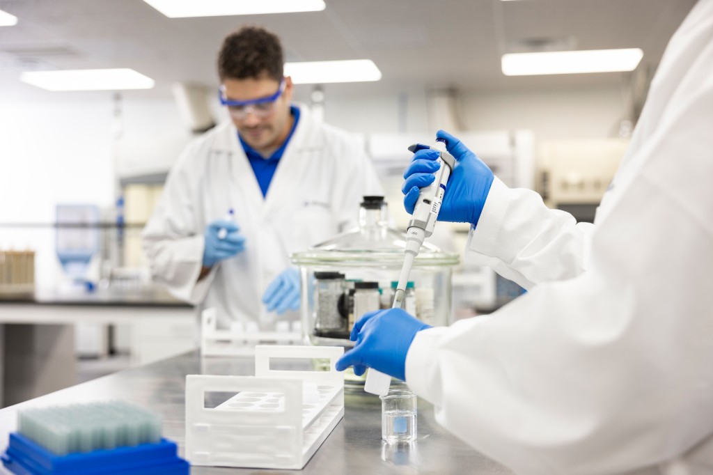 Two laboratory technicians in lab coats and safety goggles perform tests for non-intentionally added substances