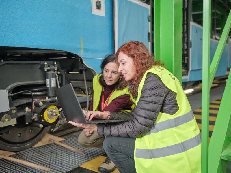Zwei Ingenieurinnen in Sicherheitswesten bei der Datenauswertung auf einem Laptop neben Schienenfahrzeugkomponenten in einer Werkstatt