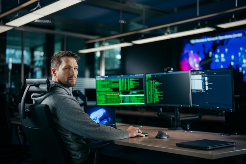 Mann an einem Schreibtisch mit mehreren Monitoren, die Programmcode anzeigen, in einem dunklen Büro