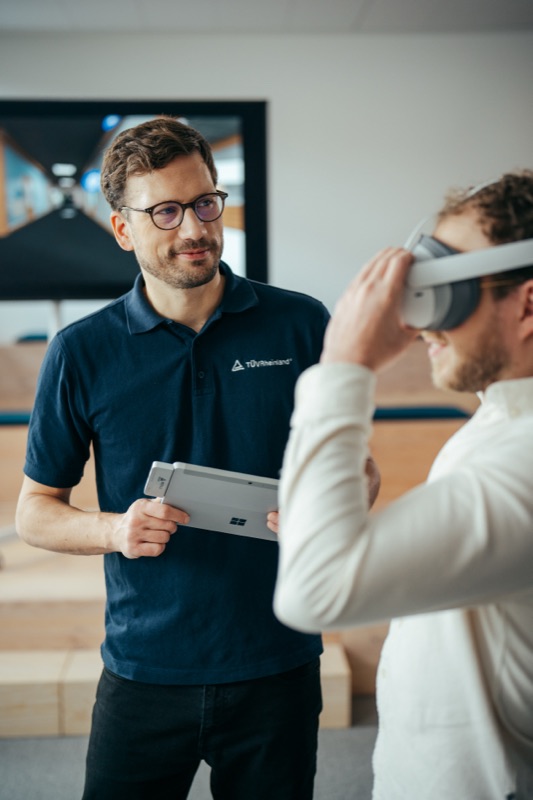 Engineer holding a tablet while a colleague adjusts a virtual reality headset in an office